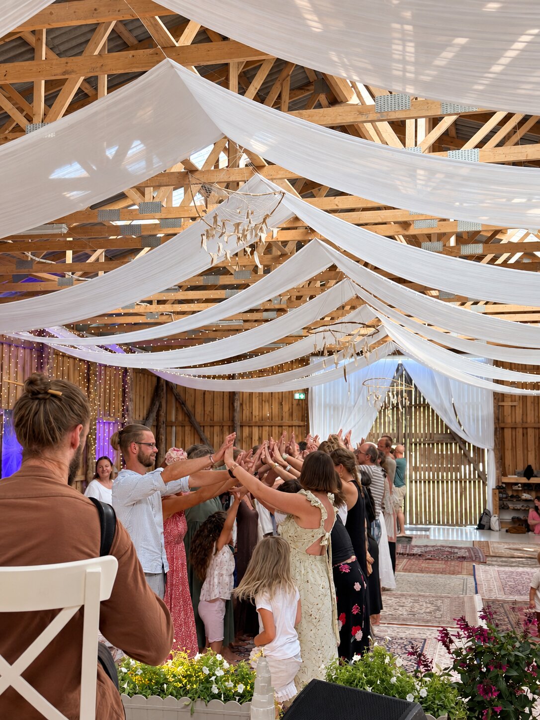 Festival participants raising their hands together in a ritual under white fabric in a wooden barn