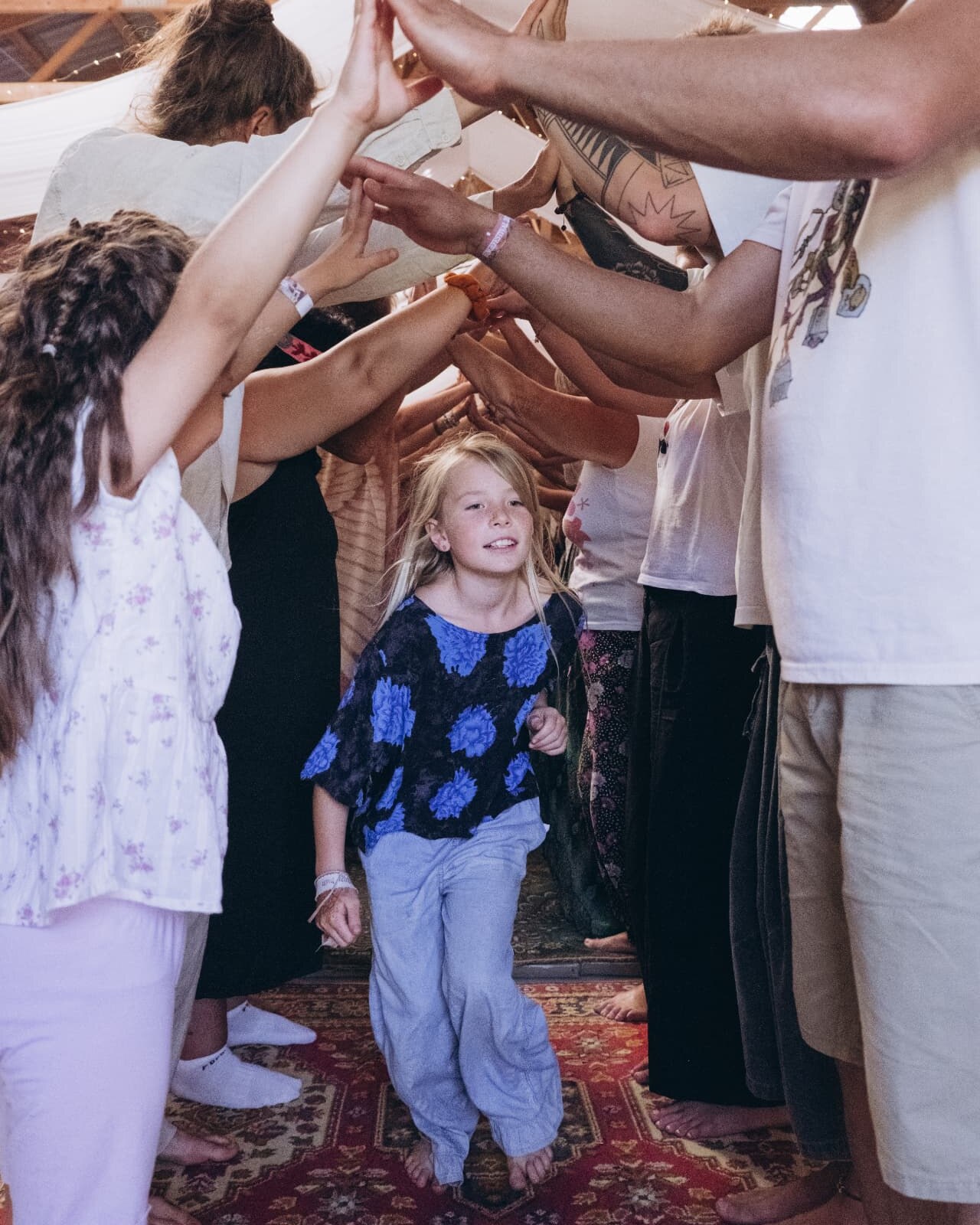 A smiling child walks barefoot through a tunnel of raised, joined hands formed by festival participants in a wooden barn