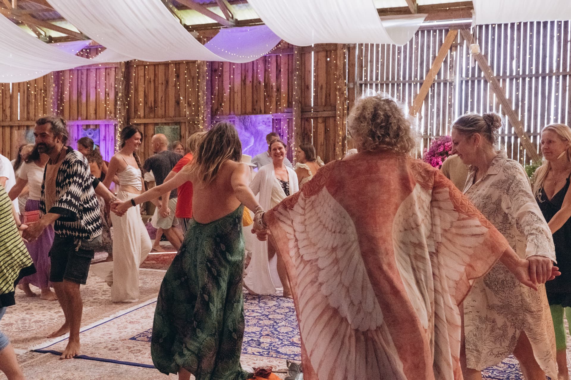Hingefestival participants holding hands in a moving circle dance on rugs in a wooden barn draped with white fabric, designed by Rituals by Joya